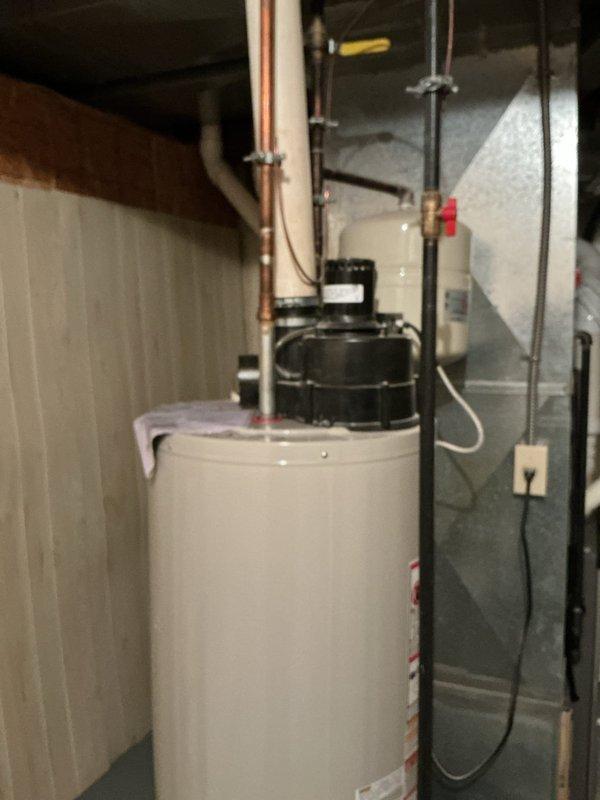 Responded to water leak service call for hot water tank. Upon inspection, found water leaking from underneath the tank in utility area. Documented current installation which includes main cylindrical water heater with connected Amtrol expansion tank, pump assembly, and associated copper/PVC piping. Leak appears to originate from tank base, requiring replacement.