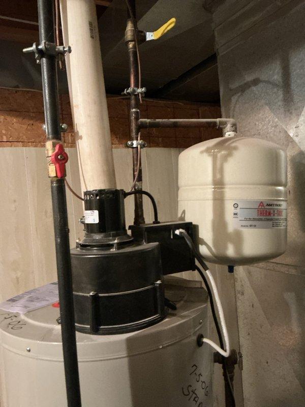 Responded to water leak service call for hot water tank. Upon inspection, found water leaking from underneath the tank in utility area. Documented current installation which includes main cylindrical water heater with connected Amtrol expansion tank, pump assembly, and associated copper/PVC piping. Leak appears to originate from tank base, requiring replacement.