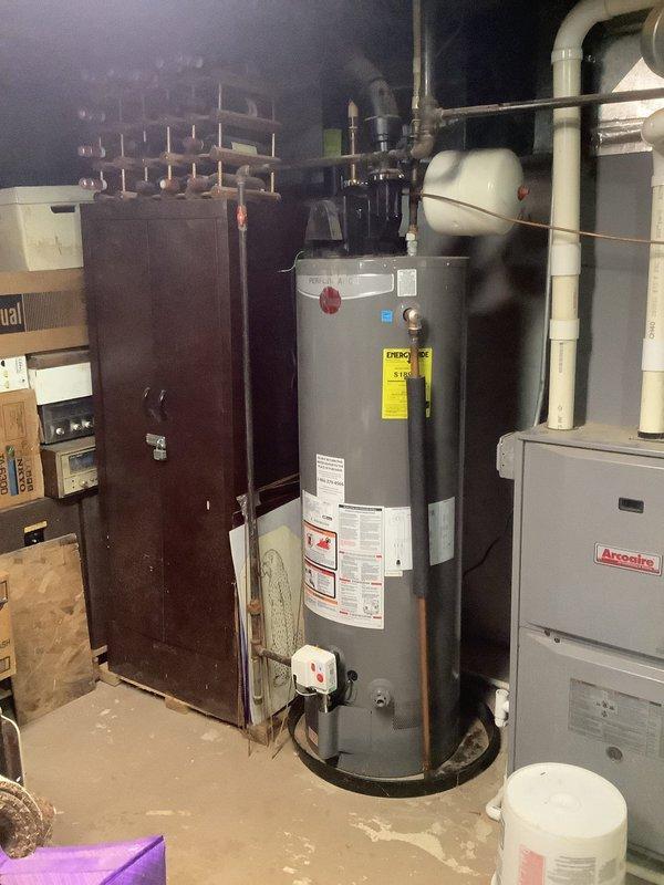 Performed diagnostic assessment on residential water heater system showing signs of wear at connection points. Inspected copper piping connections to tank and examined PVC plumbing components. Identified and repaired leaking joint at black PVC elbow pipe near the Arcoaire furnace unit, restoring proper function to the water heating system.