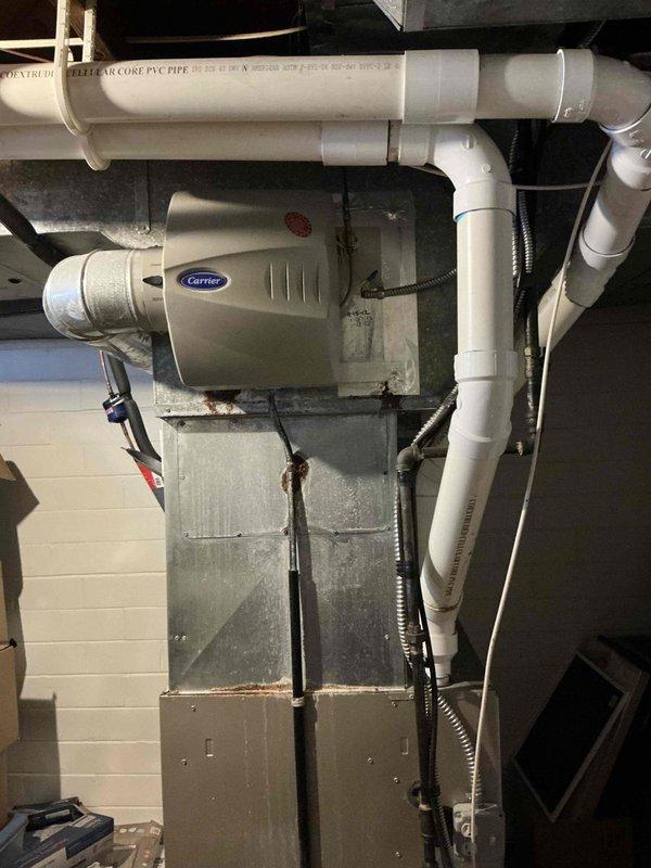 Conducted estimate for replacement of 16-year-old HVAC system. Current installation includes a Carrier humidifier mounted on the air handler with associated PVC drain lines and venting. Outdoor AC unit shows significant snow accumulation and exposure to elements, positioned against brick exterior wall with nearby drainage pipes.