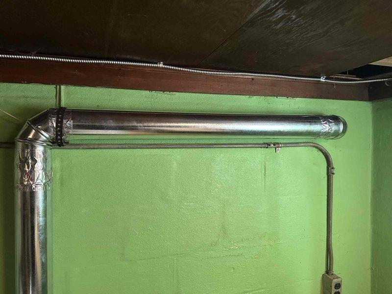 Completed installation of rigid metal dryer vent ductwork in basement laundry area, replacing non-compliant flexible hose to meet city code requirements for electric dryer ventilation. System includes properly sealed 90-degree elbow joints and secure wall mounting for optimal airflow in this 60+ year old residence.