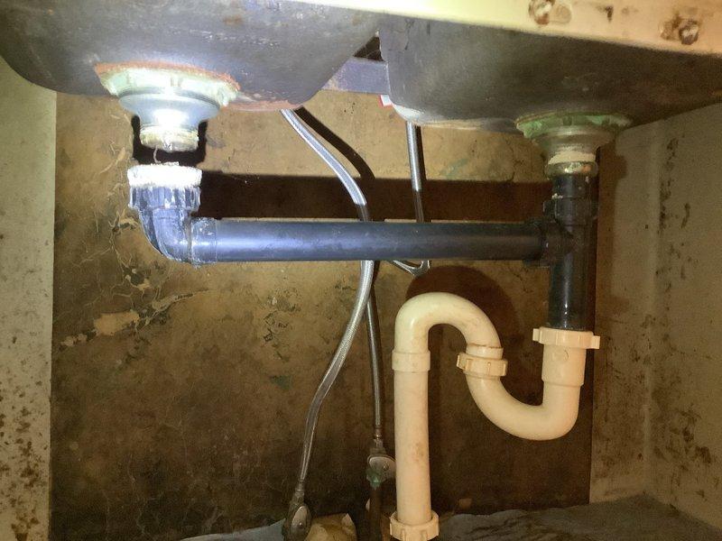 Diagnosed kitchen sink drainage system with reported leaking issues. Inspected dual-basin setup with metal sink basins connected to a gray PVC drain pipe and white P-trap. Identified mineral deposits at connection points and possible water damage on cabinet surface, indicating long-term seepage. Resolved leaking by tightening connections and ensuring proper alignment of drainage assembly.