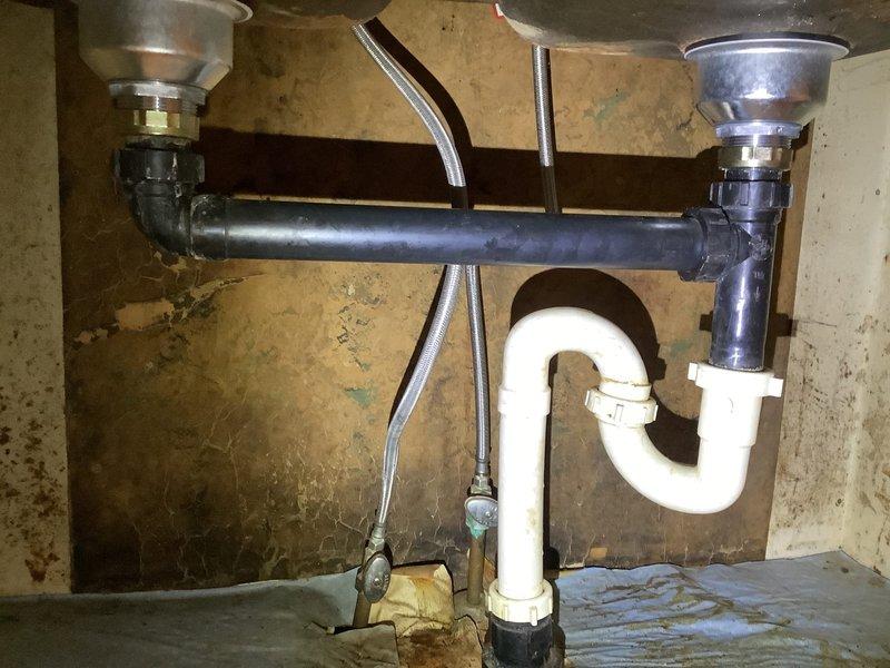 Diagnosed kitchen sink drainage system with reported leaking issues. Inspected dual-basin setup with metal sink basins connected to a gray PVC drain pipe and white P-trap. Identified mineral deposits at connection points and possible water damage on cabinet surface, indicating long-term seepage. Resolved leaking by tightening connections and ensuring proper alignment of drainage assembly.