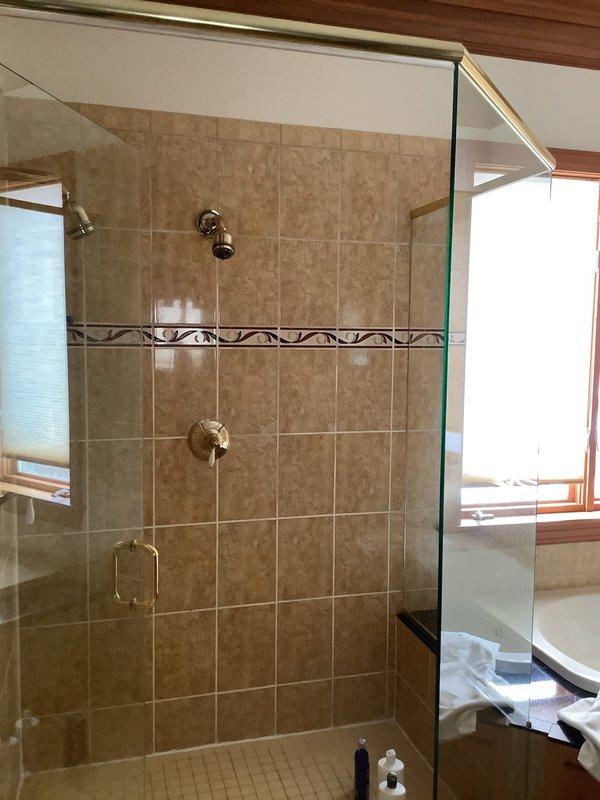 Responded to service call for leaking shower head in a glass-enclosed shower stall with beige/tan ceramic tile walls. Upon inspection, found the existing chrome shower head was malfunctioning. Removed defective unit and installed new shower head, ensuring proper sealing at connection points to prevent future leaks. Tested water flow and verified no leakage after installation.