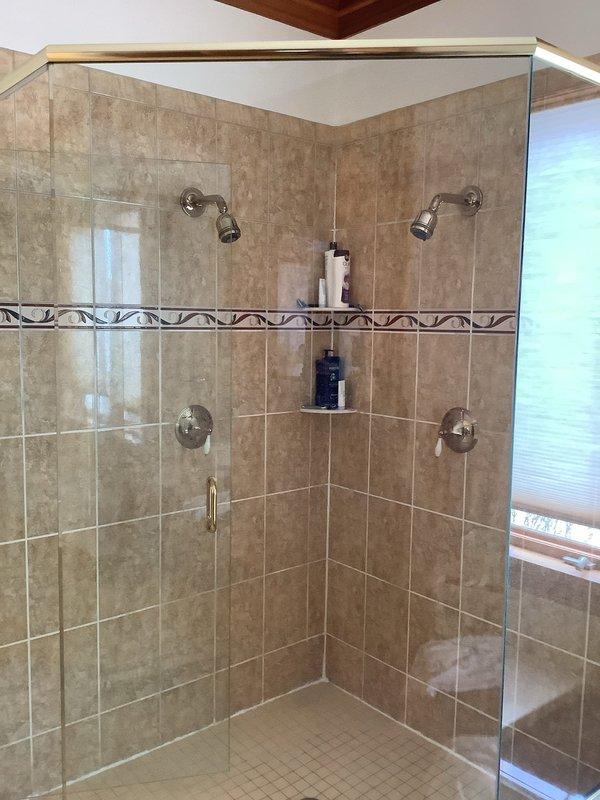 Responded to service call for leaking shower head in a glass-enclosed shower stall with beige/tan ceramic tile walls. Upon inspection, found the existing chrome shower head was malfunctioning. Removed defective unit and installed new shower head, ensuring proper sealing at connection points to prevent future leaks. Tested water flow and verified no leakage after installation.