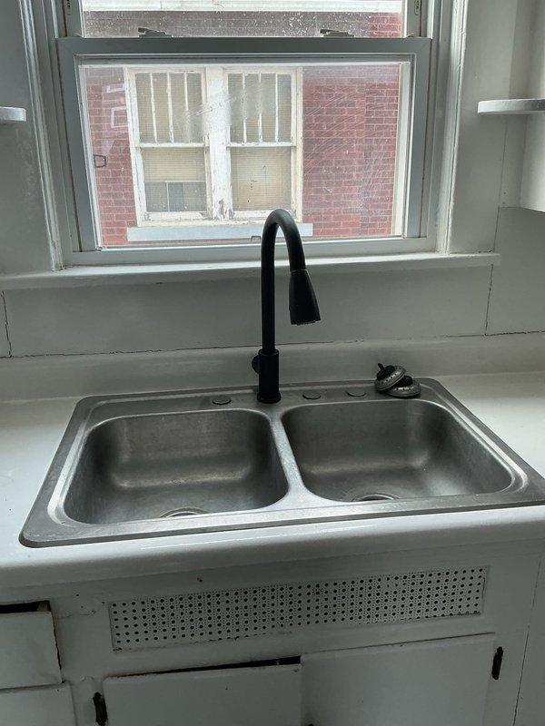 Inspected kitchen sink plumbing system with double-basin stainless steel sink and black pull-down faucet. Evaluated aged PVC P-trap assembly showing discoloration and staining, with particular attention to the white plastic coupling fittings and drain connection. Photos documented both the visible kitchen fixtures and underlying plumbing configuration for reference in inspector report.