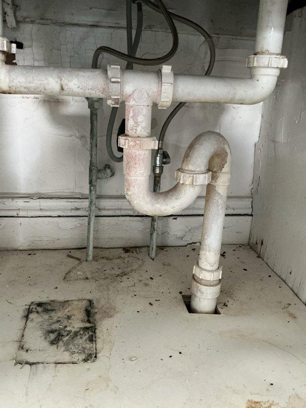 Inspected kitchen sink plumbing system with double-basin stainless steel sink and black pull-down faucet. Evaluated aged PVC P-trap assembly showing discoloration and staining, with particular attention to the white plastic coupling fittings and drain connection. Photos documented both the visible kitchen fixtures and underlying plumbing configuration for reference in inspector report.