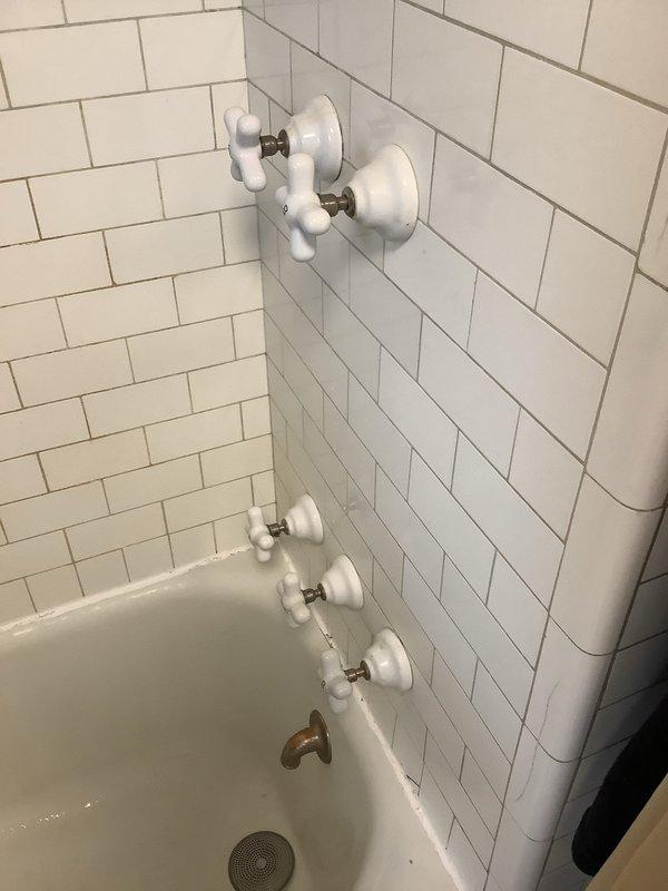Responded to service call regarding leaking plumbing stems for hot and cold water fixtures in bathroom sink. Inspected the bathroom installation featuring six porcelain cross-handle fixtures (three upper and three lower) with signs of normal wear. Further examination of the utility area revealed copper pipes with soldered joints and a central cast iron pipe with two red valve handles controlling water flow through the system.
