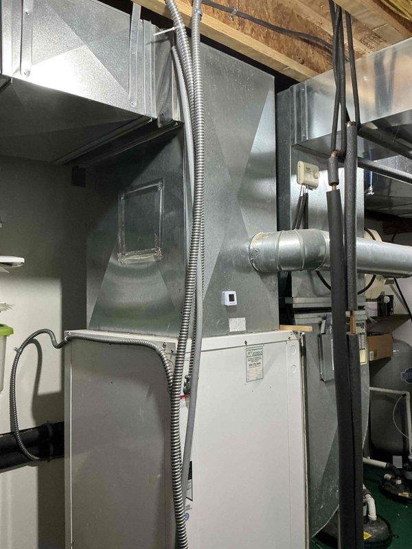 Inspected 20-year-old geothermal HVAC system in basement utility area. Documented existing installation including air handler/furnace unit with metal exhaust vent, ceiling ductwork, and associated connections for potential replacement. Photographs taken of complete system including main unit, ductwork configuration, and electrical components.