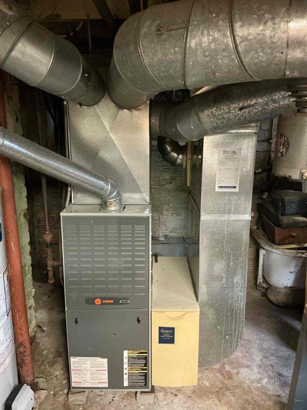 Conducted comprehensive assessment of 23-year-old furnace system with potential for AC addition as quoted. Inspection revealed weathered exterior siding with visible damage and an aging Trane furnace unit with standard galvanized ductwork in basement. Documented existing setup including connected humidifier/air purifier unit to provide replacement options for complete HVAC system upgrade.