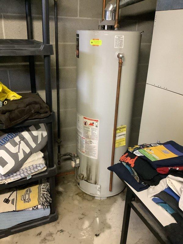 Performed inspection of 26-year-old 50-gallon gas water heater exhibiting significant leakage throughout unit. Photos documented aged unit installed against basement cinder block wall with copper supply lines and metal vent piping. Provided estimate for complete replacement system including new water heater, required code-compliant connections, and proper disposal of existing unit.