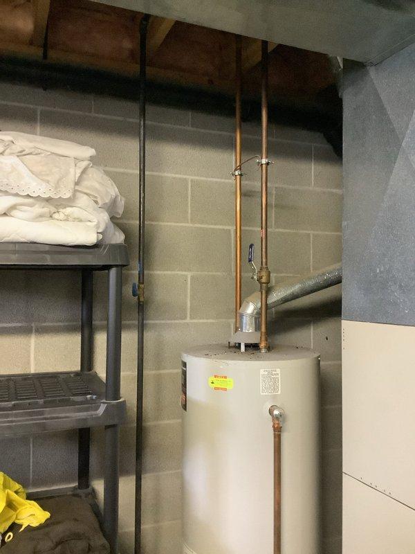 Performed inspection of 26-year-old 50-gallon gas water heater exhibiting significant leakage throughout unit. Photos documented aged unit installed against basement cinder block wall with copper supply lines and metal vent piping. Provided estimate for complete replacement system including new water heater, required code-compliant connections, and proper disposal of existing unit.