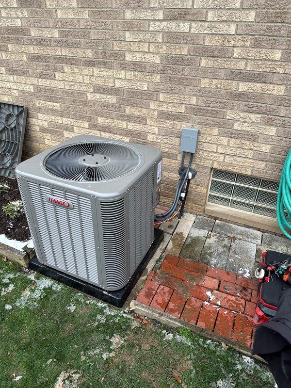 Installed new 90K BTU Lennox air conditioning system with outdoor condenser unit properly mounted on code-compliant pad adjacent to home exterior. Completed electrical connections via wall-mounted disconnect box with appropriate conduit and power lines. System was tested and verified to be functioning according to manufacturer specifications.