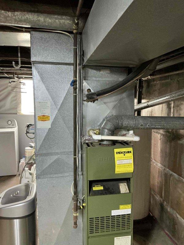 Conducted inspection of 1984 Rheem furnace identified as winner of Oldest Furnace Giveaway program. Unit is olive-green with visible warning labels, connected to gray ductwork with 90-degree bend extending into wall. Documented current installation including surrounding infrastructure (copper piping, concrete block walls) to prepare for upcoming replacement. Left message to confirm details for the scheduled replacement.
