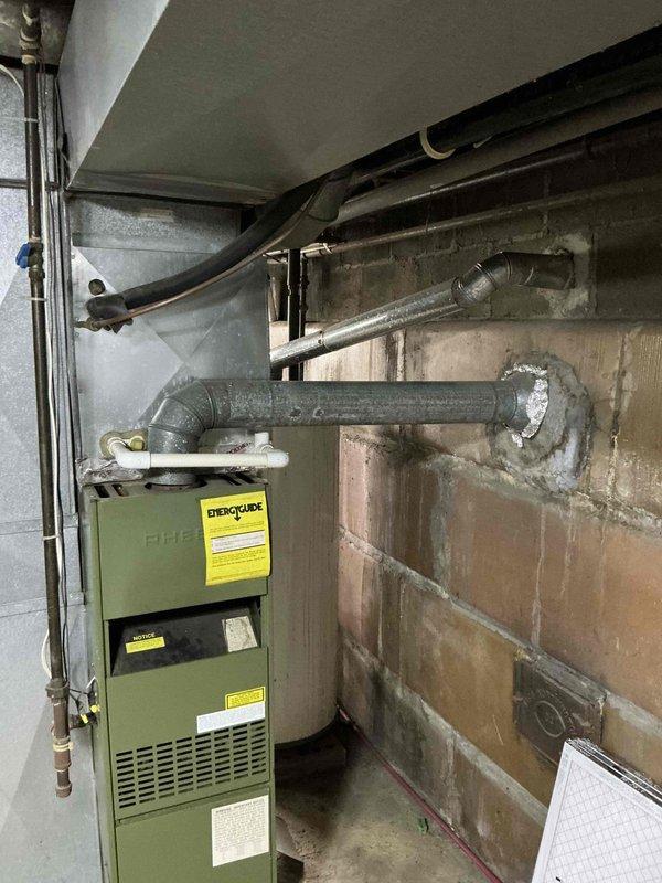 Conducted inspection of 1984 Rheem furnace identified as winner of Oldest Furnace Giveaway program. Unit is olive-green with visible warning labels, connected to gray ductwork with 90-degree bend extending into wall. Documented current installation including surrounding infrastructure (copper piping, concrete block walls) to prepare for upcoming replacement. Left message to confirm details for the scheduled replacement.