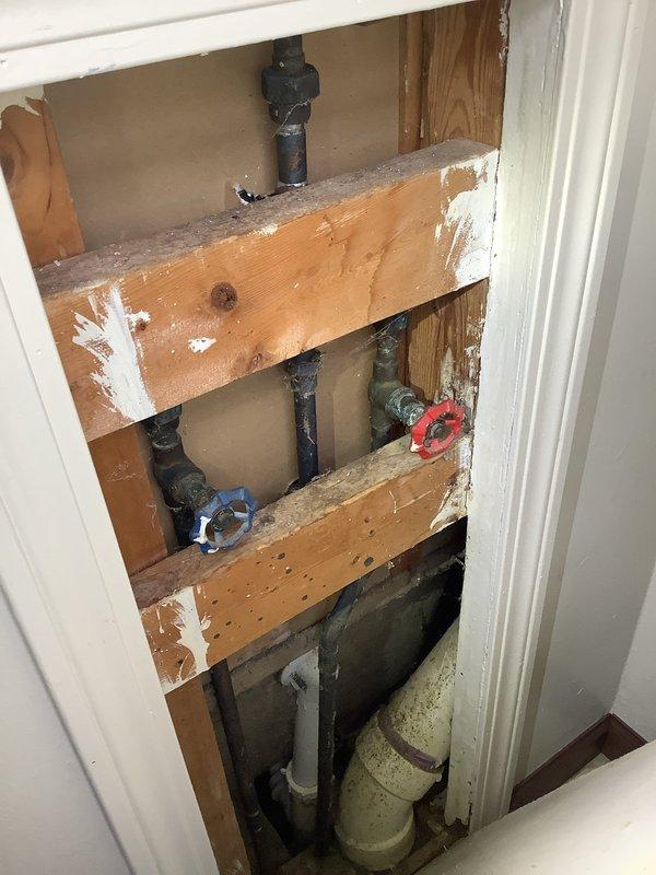 # Plumbing Service Report

Responded to service call for plumbing issue in 40-year-old residential property. Upon arrival, performed comprehensive inspection of affected plumbing systems, including under-sink connections and wall cavity piping.

Inspection revealed concerns with aging pipe infrastructure requiring access through interior wall cavity. Removed section of finished wall to expose plumbing chase containing vertical supply lines, drain stacks, and shut-off valves. Documented existing conditions showing cast iron/black steel piping, PVC drain lines, and braided stainless steel flexible supply connections beneath bathroom sink fixture.

Assessed P-trap assembly, drain connections, and water supply lines for leaks, corrosion, or damage. Verified proper operation of red and blue-handled shut-off valves controlling water supply to affected fixtures. Examined structural wooden framing for water damage or deterioration from potential previous leaks.

Completed necessary repairs to 