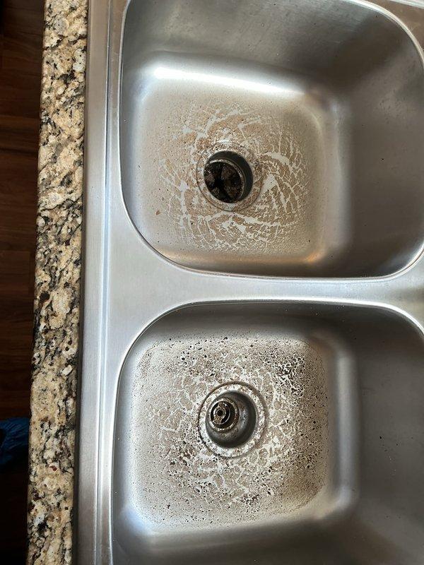 Responded to service call for clogged kitchen sink in 20-year-old residence. Upon arrival, inspected stainless steel double-bowl sink with visible standing water and soap residue in both basins indicating drainage issues. Accessed cabinet beneath sink and examined plumbing assembly including P-trap, drain lines, and supply connections. Cleared blockage from drain system restoring proper water flow through both sink bowls. Verified P-trap and all PVC connections were secure with no leaks present. Tested drainage by running water through both basins to confirm complete resolution of clog. All plumbing components left in proper working condition.