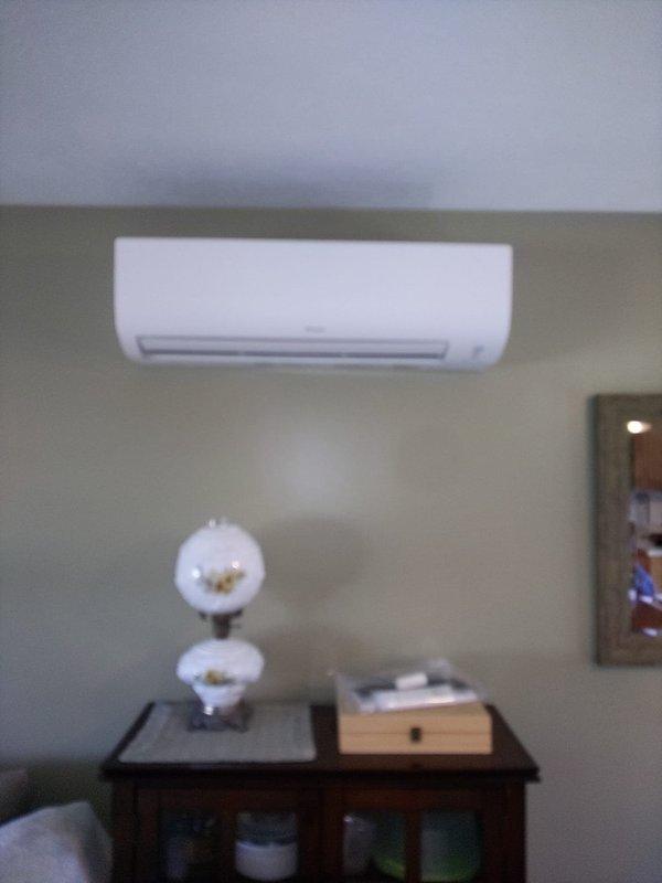 Completed final installation phase of ductless mini-split air conditioning system by concealing refrigerant line set and electrical conduit. Installed exterior line hide cover along building exterior wall, securing two vertical white PVC chase sections to conceal refrigerant lines running from ground level to the wall-mounted indoor air handler unit, ensuring professional appearance and protection of refrigerant piping from environmental elements.