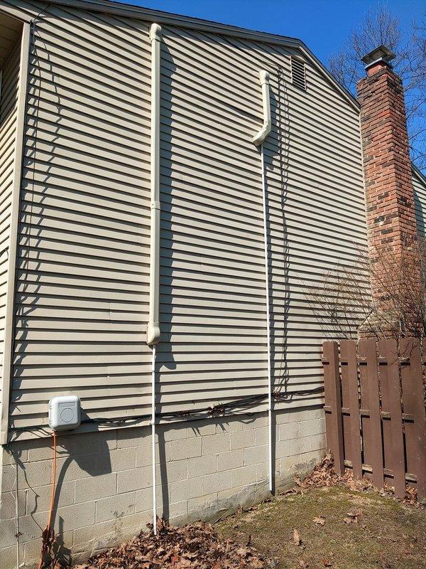 Completed final installation phase of ductless mini-split air conditioning system by concealing refrigerant line set and electrical conduit. Installed exterior line hide cover along building exterior wall, securing two vertical white PVC chase sections to conceal refrigerant lines running from ground level to the wall-mounted indoor air handler unit, ensuring professional appearance and protection of refrigerant piping from environmental elements.