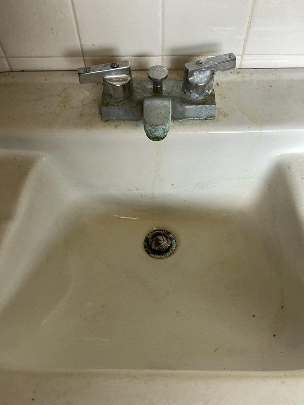Responded to a multi-issue plumbing service call at a 60-year-old residential property addressing clogged drain, water leak, and toilet repair. Upon arrival, inspected utility sink with significant mineral buildup and staining around drain assembly and faucet fixtures. Cleared obstruction from drain line using appropriate tools and verified proper water flow. Examined plumbing components in confined access area beneath sink, identifying deteriorated conditions including rust staining, water damage, and corrosion on surrounding surfaces. Inspected white PVC piping connections and joints for integrity, noting the presence of a blue valve assembly in the system. Addressed leak source by tightening connections and assessing condition of aging pipes showing signs of prolonged moisture exposure. Performed toilet repair by examining tank components and making necessary adjustments to restore proper function. Tested all fixtures following repairs to ensure proper operation and drainage. Docume