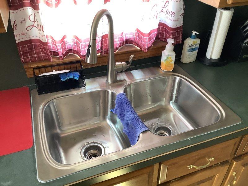 Removed existing kitchen sink and faucet assembly, then installed a new Kohler double-basin stainless steel sink with Moen Arbor spot-resistant pull-down faucet. Installation included new chrome basket strainer assemblies for both basins and complete rework of the drain system underneath, configuring black PVC P-trap assembly to properly accommodate the new double-basin setup. Cleaned and organized the cabinet interior upon completion, ensuring all plumbing connections were secure and leak-free.