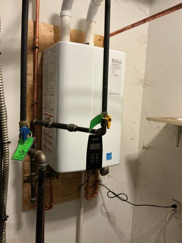 Completed professional installation of a Navien tankless water heater system in a basement utility area. The installation included mounting the unit on a plywood backing board, connecting copper supply and return lines with shutoff valves, and installing dual PVC venting pipes through the ceiling. All plumbing connections were properly fitted with appropriate valves and fittings, and the system was tested for proper operation. The installation meets manufacturer specifications and energy efficiency standards as indicated by Energy Star certification.