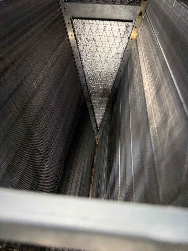 Completed scheduled BS-MVP heating maintenance visit and performed comprehensive system inspection. Documented evaporator coil condition showing significant frost accumulation on heat exchanger fins, indicating active refrigeration cycle operation. Photographed interior components including corrugated metal housing panels and coil assembly within confined vertical channel space to verify proper system operation and identify any maintenance concerns.