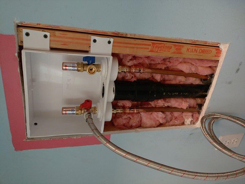 Replaced washer box in first-level laundry room by cutting open wall access to reach plumbing infrastructure behind the installation area. Connected new supply lines to existing drain line and installed new shut-off valves with brass fittings, including one valve with blue handle and one with red handle, mounted on white plastic mounting plate secured to wall studs. Installed customer-supplied braided metal washer hose lines to replace existing connections. Wall cavity work exposed fiberglass insulation between studs, with all plumbing connections completed and tested for proper operation. Drywall repair left for homeowner responsibility as agreed.