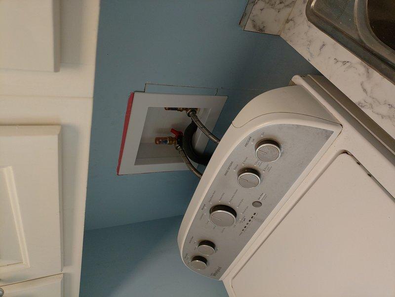 Replaced washer box in first-level laundry room by cutting open wall access to reach plumbing infrastructure behind the installation area. Connected new supply lines to existing drain line and installed new shut-off valves with brass fittings, including one valve with blue handle and one with red handle, mounted on white plastic mounting plate secured to wall studs. Installed customer-supplied braided metal washer hose lines to replace existing connections. Wall cavity work exposed fiberglass insulation between studs, with all plumbing connections completed and tested for proper operation. Drywall repair left for homeowner responsibility as agreed.