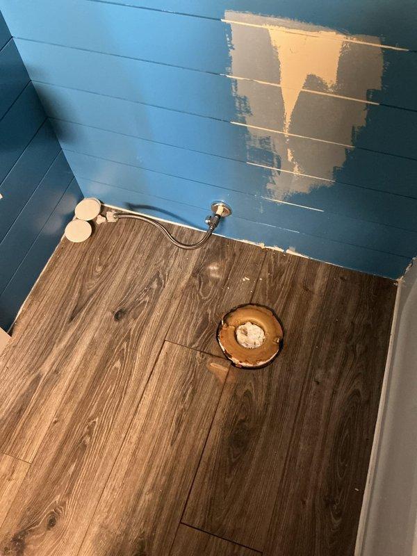 Completed full toilet installation in mobile home bathroom. Removed existing toilet flange and prepared subfloor opening, installed new wax ring and closet flange, and set new two-piece white toilet with chrome flush lever. Connected water supply line to shut-off valve, secured toilet to floor with mounting bolts, tested flush operation and checked for leaks. Bathroom features laminate wood-look flooring and teal shiplap walls in compact layout adjacent to bathtub/shower enclosure.