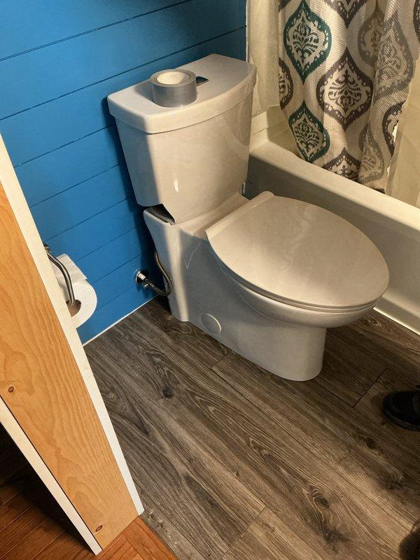 Completed full toilet installation in mobile home bathroom. Removed existing toilet flange and prepared subfloor opening, installed new wax ring and closet flange, and set new two-piece white toilet with chrome flush lever. Connected water supply line to shut-off valve, secured toilet to floor with mounting bolts, tested flush operation and checked for leaks. Bathroom features laminate wood-look flooring and teal shiplap walls in compact layout adjacent to bathtub/shower enclosure.