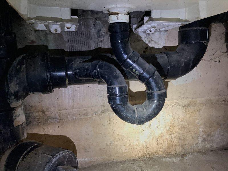 Conducted main line repair estimate for stack replacement in basement utility area. Assessed existing vertical drainage pipes running along cinder block wall and evaluated P-trap configuration beneath sink fixture. Documented current plumbing infrastructure conditions including aged black drain pipes, mounting hardware, and wall-mounted drain assembly showing signs of moisture exposure and typical wear. Technician to return following completion of restoration company's work to finalize stack replacement installation.