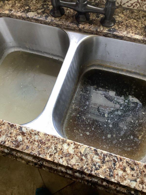 Responded to clogged kitchen sink in 1960s-era home. Upon arrival, found double-basin stainless steel sink with standing water in both basins—left basin contained relatively clear water while right basin had dark, murky water with visible sediment and debris accumulation. Inspected under-sink plumbing configuration which included white PVC P-trap assembly connected to garbage disposal unit. Cleared blockage from drain line, restored proper drainage to both basins, and verified garbage disposal operation. System tested and functioning normally upon completion.