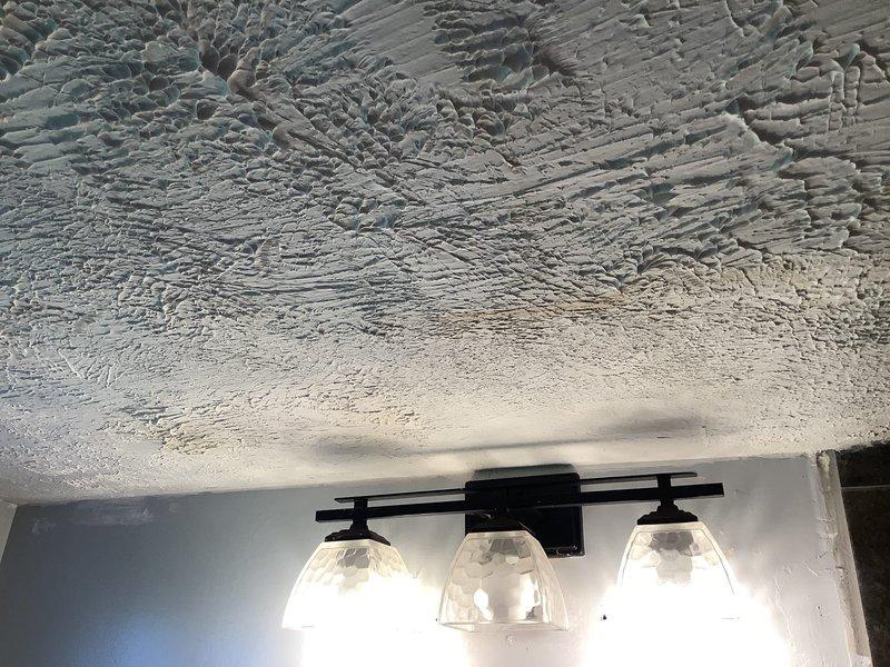 Responded to report of suspected leak originating from upstairs bathroom tub area with visible water damage on downstairs bathroom ceiling. Performed visual inspection and documentation of affected ceiling area, which displayed water staining and damage to popcorn texture finish near bathroom vanity light fixture. Utilized camera equipment to photograph and document extent of ceiling damage for diagnostic purposes. Evidence of active or previous water intrusion was confirmed through visual assessment of compromised ceiling material.