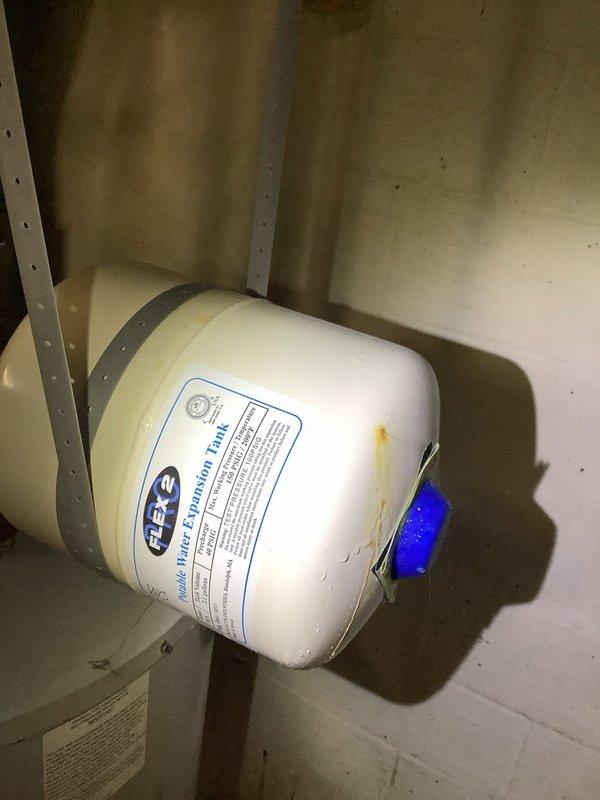 Responded to a water leak in basement originating from the potable water expansion tank. Upon arrival, found a Flexcon water expansion tank with a minor leak at the connection point, causing spray and moisture on surrounding walls and floor. Inspected the white cylindrical expansion tank mounted in the utility area near the water heater and furnace system. The leak was contained to light spray without significant pooling. Assessed copper piping connections to the expansion tank and evaluated the overall condition of the unit, which showed signs of age with visible discoloration and staining on the exterior surface. Documented the leak location at the blue valve assembly connection point on the tank. The expansion tank was positioned horizontally in the basement utility room alongside the main water heater and HVAC equipment.