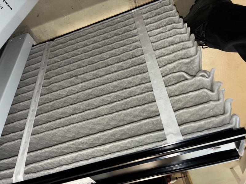 Completed scheduled MVP cooling maintenance visit. Inspected and documented HVAC air filtration system, noting moderate dust and particulate accumulation on pleated air filter within the air handler unit. Filter media showed typical service wear with visible debris collection across the surface, indicating normal system operation and adequate particle capture. Photographed filter condition for maintenance records while installed in black plastic housing frame mounted on outdoor decking area.