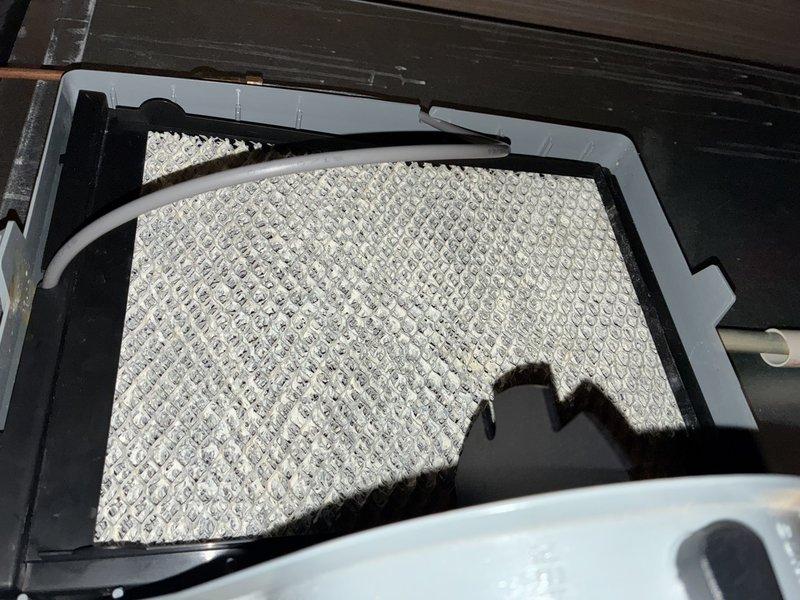 Completed scheduled MVP cooling maintenance visit. Inspected and documented HVAC air filtration system, noting moderate dust and particulate accumulation on pleated air filter within the air handler unit. Filter media showed typical service wear with visible debris collection across the surface, indicating normal system operation and adequate particle capture. Photographed filter condition for maintenance records while installed in black plastic housing frame mounted on outdoor decking area.
