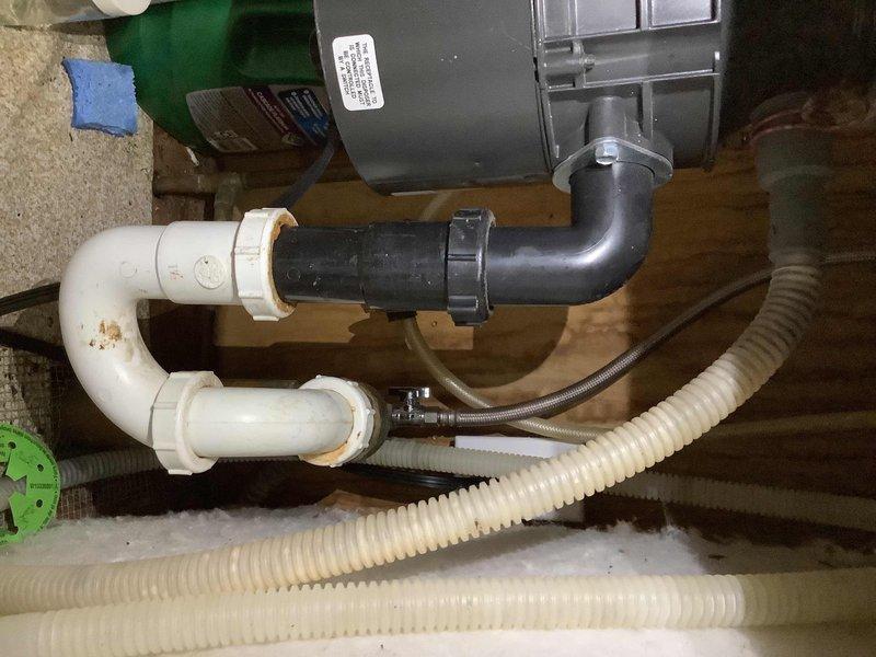 Responded to water leak complaint originating from dishwasher and kitchen sink area causing water damage to basement ceiling and floor. Upon inspection, identified compromised plumbing connections in the drainage system serving the kitchen fixtures. Located source of leak at dishwasher drain line connection point where corroded PVC piping and deteriorated trap assembly were allowing water to escape during drainage cycles. 

Documented existing conditions showing rust-stained white PVC trap configuration with signs of age-related deterioration and mineral buildup at pipe joints. Noted flexible discharge hoses and corrugated drain lines connected to gray pump assembly, which services the dishwasher drainage system. Inspected ceiling access panel revealing water intrusion path through building cavity affecting insulation and structural framing members.

Photographed damaged areas including compromised fire-rated mesh material in ceiling access opening and affected mechanical equipment hou