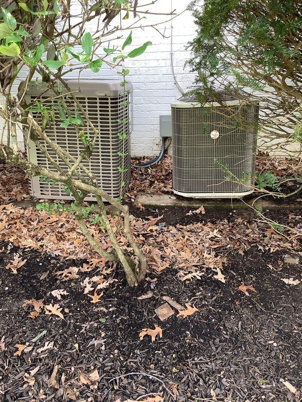 Completed preventative maintenance tune-ups on two outdoor condensing units at residential property. The older unit on the left showed significant vegetation encroachment with plant growth penetrating the protective grille and heavy debris accumulation on condenser coils, while the newer cylindrical unit on the right was in better overall condition with less environmental impact. Performed standard maintenance procedures including cleaning condenser coils, removing organic debris from both units, and clearing vegetation from around equipment to ensure proper airflow and system efficiency.