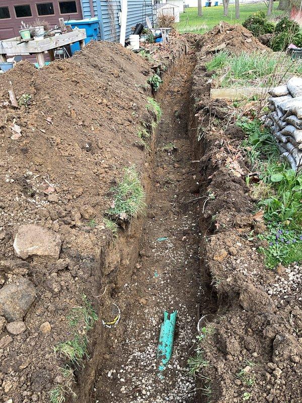 Excavated approximately 80 linear feet of trench from rear of residence to front right side of driveway to remove and replace deteriorated clay tile sewer line with broken sections and bellied areas. Trench averaged 2-3 feet in width and several feet in depth to achieve proper pitch. Removed existing cast iron pipe penetrating basement wall and replaced with new PVC piping to establish correct drainage gradient. Installation included new white PVC sewer line running full length of excavation. Utilized 10-foot excavator for digging operations. Backfilled trench with excavated material and topped with 1.5 yards of #57 limestone, mounding soil above grade to allow for settling. Removed small trees and bushes as necessary to complete excavation work. Two-person crew completed project in approximately 8 hours.