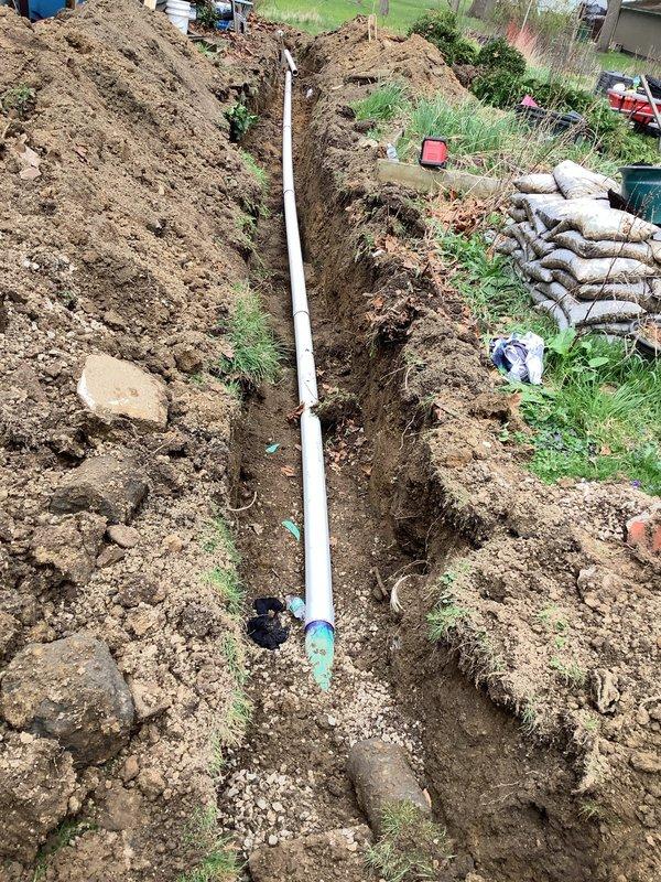 Excavated approximately 80 linear feet of trench from rear of residence to front right side of driveway to remove and replace deteriorated clay tile sewer line with broken sections and bellied areas. Trench averaged 2-3 feet in width and several feet in depth to achieve proper pitch. Removed existing cast iron pipe penetrating basement wall and replaced with new PVC piping to establish correct drainage gradient. Installation included new white PVC sewer line running full length of excavation. Utilized 10-foot excavator for digging operations. Backfilled trench with excavated material and topped with 1.5 yards of #57 limestone, mounding soil above grade to allow for settling. Removed small trees and bushes as necessary to complete excavation work. Two-person crew completed project in approximately 8 hours.