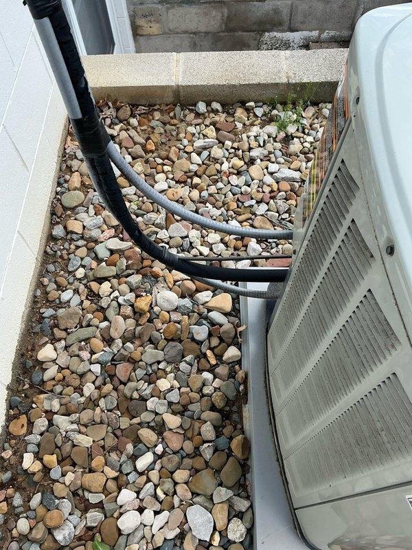 Conducted on-site assessment for complete HVAC system replacement, evaluating existing Trane air conditioning condenser unit and associated components. Documented current equipment condition, noting weathered exterior surfaces, existing refrigerant line routing with black foam insulation, electrical conduit configuration, and outdoor installation layout on concrete pad with rock landscaping. Photographed equipment specifications, mounting platform, wall-mounted electrical components, and accessibility for proposed furnace and air conditioning system replacement.