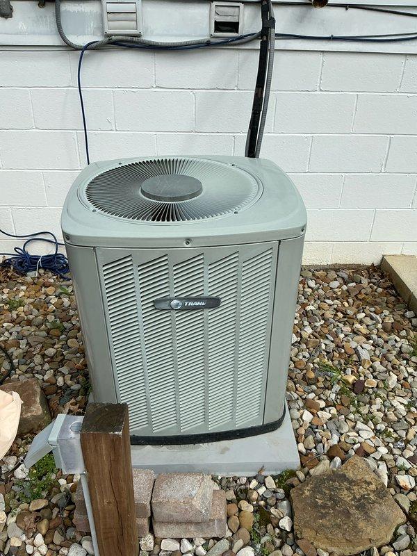 Conducted on-site assessment for complete HVAC system replacement, evaluating existing Trane air conditioning condenser unit and associated components. Documented current equipment condition, noting weathered exterior surfaces, existing refrigerant line routing with black foam insulation, electrical conduit configuration, and outdoor installation layout on concrete pad with rock landscaping. Photographed equipment specifications, mounting platform, wall-mounted electrical components, and accessibility for proposed furnace and air conditioning system replacement.