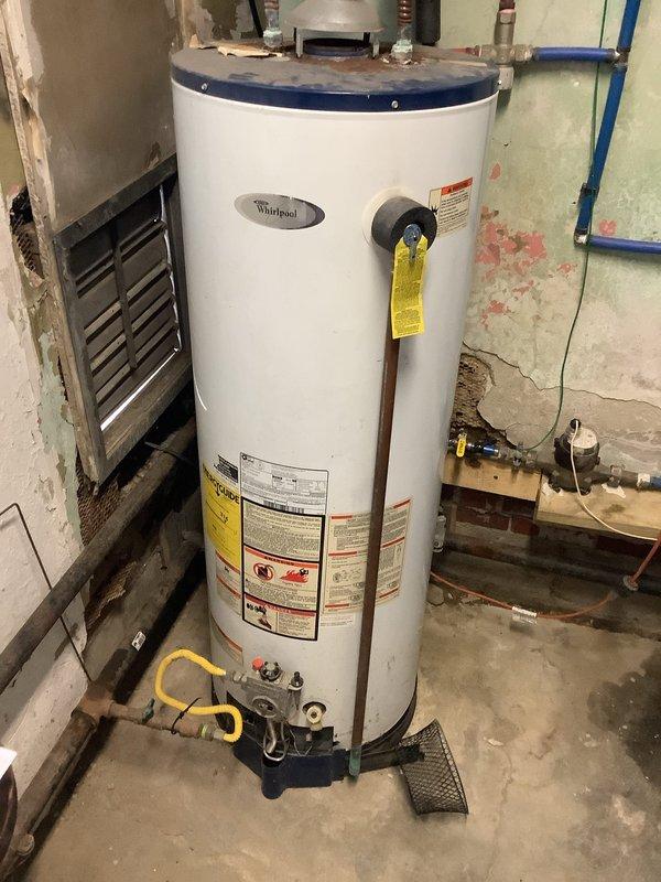 Responded to service call for Whirlpool tanked water heater that had been red-tagged by gas utility company due to missing sediment trap on gas line. Upon arrival, confirmed unit was shut down and tagged out of service for safety code violation. Inspected gas connection and plumbing configuration, verified absence of required sediment trap per code requirements. Customer also reported no hot water production prior to red tag issuance. Documented existing conditions including yellow flexible gas line connection, copper supply piping, and overall installation setup for code compliance assessment and repair recommendations.