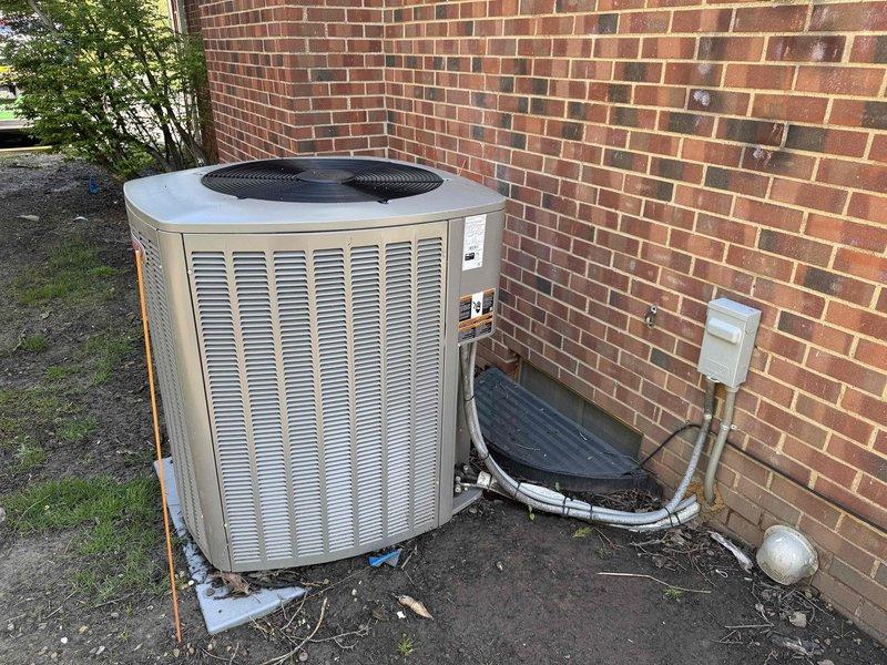 Completed BS-MVP recurring cooling maintenance visit on outdoor condensing unit. Performed visual inspection of beige/tan air conditioning condenser positioned on concrete pad against brick wall. Unit accessible with electrical disconnect box properly mounted. Documented current condition of heat exchanger coils, noting moderate to heavy accumulation of dirt, debris, and environmental soiling on condenser fin surfaces. Left section of coil exhibited brownish-tan deposits and staining requiring attention. Right section showed vertical streaking and darker patches from accumulated particulate matter. Verified unit operational status and confirmed all manufacturer specification tags present and legible. Photographed equipment condition including louvered side panels, black fan grille cover, refrigerant line set, and condensate management components for maintenance records. Unit foundation stable with minimal debris around perimeter.