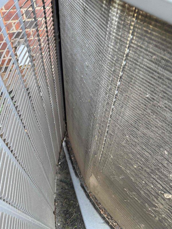 Completed BS-MVP recurring cooling maintenance visit on outdoor condensing unit. Performed visual inspection of beige/tan air conditioning condenser positioned on concrete pad against brick wall. Unit accessible with electrical disconnect box properly mounted. Documented current condition of heat exchanger coils, noting moderate to heavy accumulation of dirt, debris, and environmental soiling on condenser fin surfaces. Left section of coil exhibited brownish-tan deposits and staining requiring attention. Right section showed vertical streaking and darker patches from accumulated particulate matter. Verified unit operational status and confirmed all manufacturer specification tags present and legible. Photographed equipment condition including louvered side panels, black fan grille cover, refrigerant line set, and condensate management components for maintenance records. Unit foundation stable with minimal debris around perimeter.