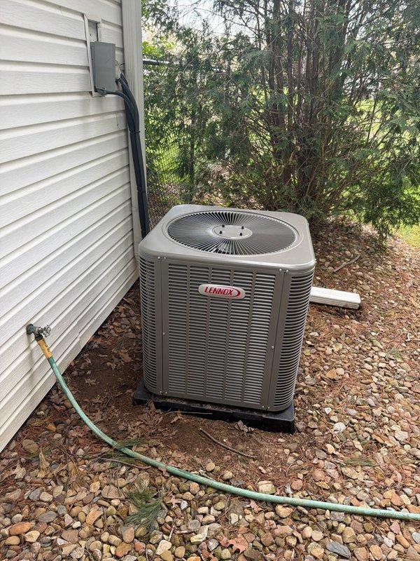 Completed installation of Lennox air conditioning condenser unit and evaporator coil system at residential property. Unit was positioned on rubber isolation pad with proper gravel base for drainage and stability, mounted adjacent to structure with white vinyl siding. Installed and routed refrigerant line set with insulated gray covering, connected copper refrigerant piping with appropriate fittings and isolation valves, and ran electrical conduit from disconnect box to unit. Secured all piping and conduit runs along exterior wall, ensured proper refrigerant connections with brass service valves, and verified all electrical connections met code requirements. System tested and operational upon completion.