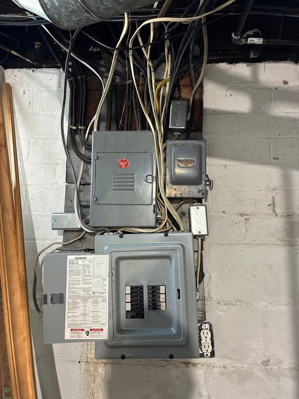 Responded to service call for electrical panel evaluation and estimate preparation. Upon arrival, inspected existing electrical service installation consisting of main breaker panel mounted on basement wall with associated junction boxes and outdated fuse panel above. Main panel appeared to be in serviceable condition with visible circuit breakers and directory labeling, though surrounding infrastructure showed age-related wear. 

Documented older fuse-style panel with screw-in fuses and brass terminal connections, noting multiple colored conductors entering the enclosure. Observed mixed vintage wiring throughout the installation with tan-colored conduits routing to ceiling area. Assessed overall electrical system configuration including panel capacity, existing circuit distribution, and connection points.

Photographed current electrical service setup to support estimate preparation for new breaker panel installation as requested. Provided customer with documentation of existing condi