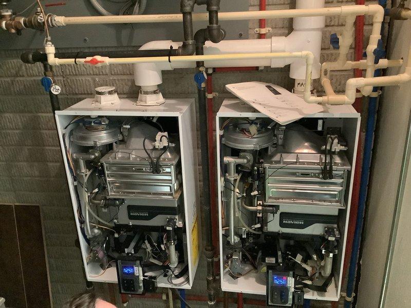 Responded to service call for non-operational tankless water heater system in single-family residence. Upon arrival, inspected dual Navien tankless water heater installation with units mounted side-by-side. Removed front access panels to assess internal components and diagnose malfunction. Observed excessive condensation accumulation on internal black plastic components and housing, indicating potential drainage or ventilation issues. Verified digital display functionality showing temperature reading of 125°F on operational unit. Examined heat exchanger assemblies, burner components, control boards, and circuit systems for both units. Inspected associated plumbing connections including PVC venting, copper supply lines, and isolation valves with proper hot/cold identification. Evaluated electrical connections and wiring integrity throughout both systems. Addressed the reported operational failure and restored system functionality. Documented existing installation configuration and inter