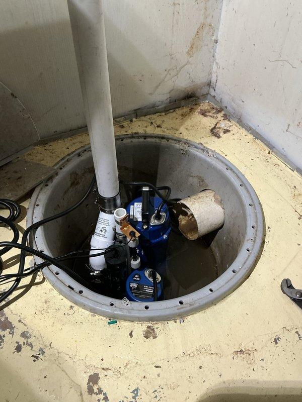 Replaced battery backup system for sump pump installation in basement utility area. New blue submersible pump installed in 18-24 inch concrete basin with white PVC discharge piping, black electrical components, and wall-mounted control switch. Parts received and system fully operational with backup power protection in place.
